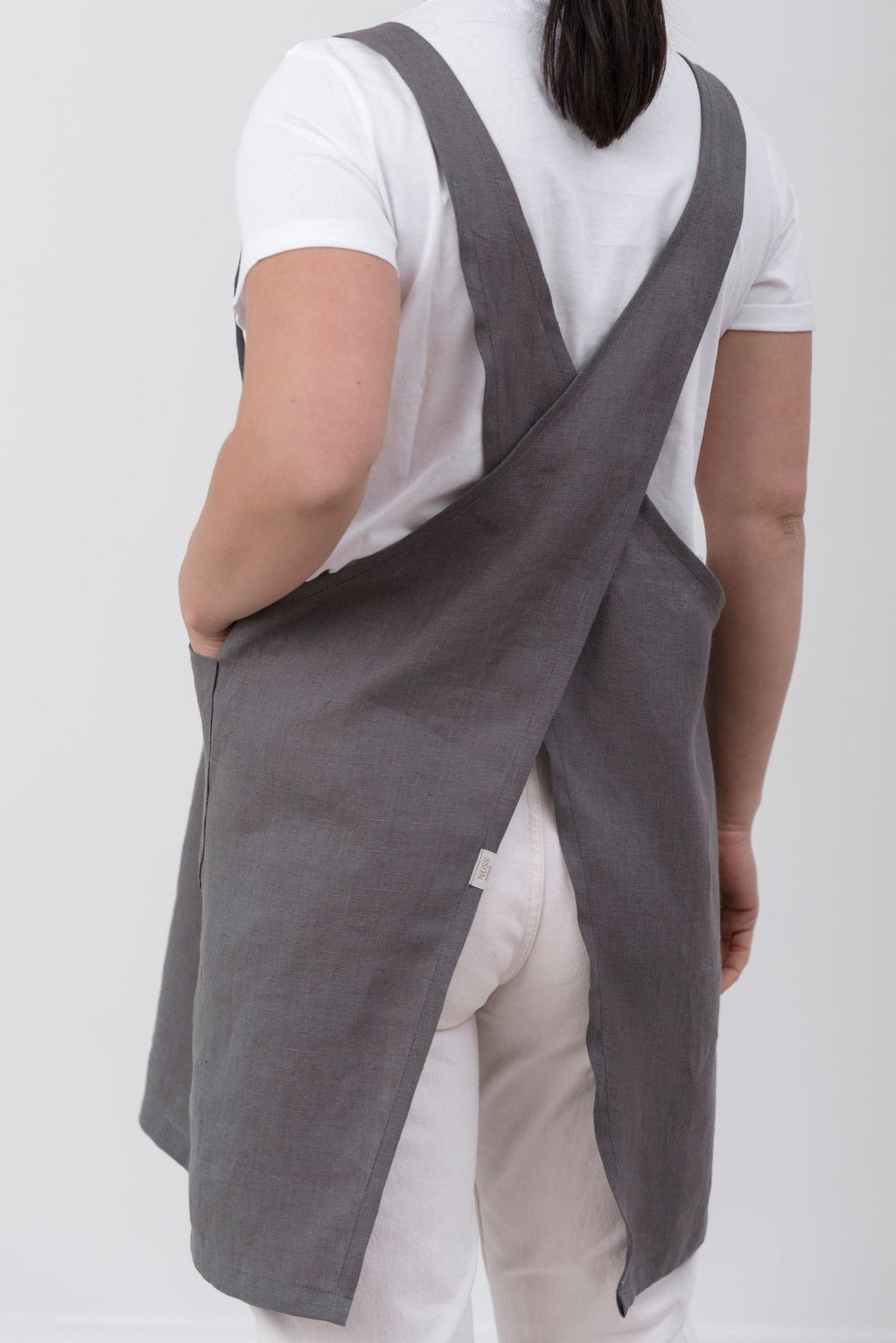 13. Woman wearing grey Japanese-style linen apron with cross-back design, shown from back, over white shirt and pants