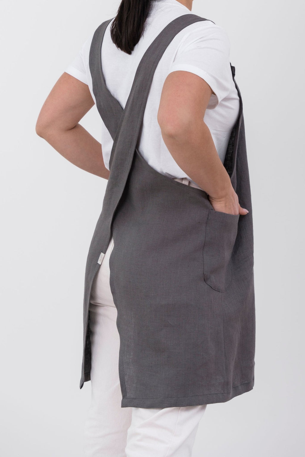 1. Woman wearing grey Japanese-style linen apron with cross-back design, shown from back, over white shirt and pants
