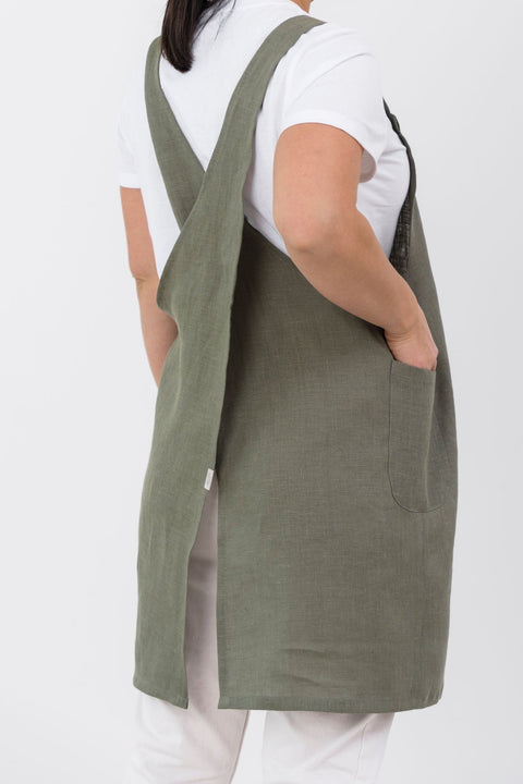 2. Woman wearing olive green Japanese-style linen apron with cross-back design, shown from back, over white shirt and pants