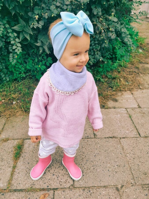 2. Child in light blue muslin scarf-bib and headband with pink sweater and boots smiling on a garden path
