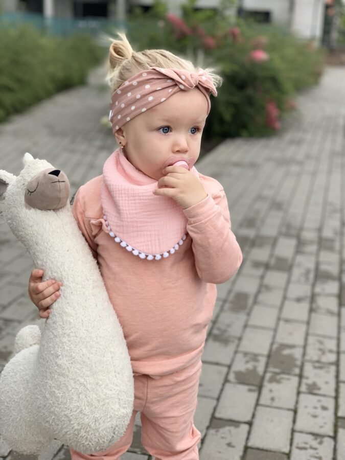 3. Close-up of child in light pink muslin scarf-bib and polka dot headband holding plush toy