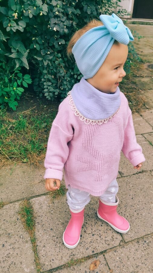 1. Child wearing light blue muslin scarf-bib and matching headband with pink sweater and boots standing on a garden path