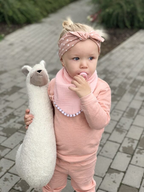 2. Child in light pink muslin scarf-bib and polka dot headband holding plush toy on paved path