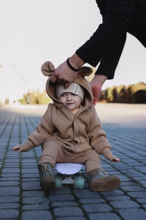 1. Baby sitting on skateboard wearing cacao eared jumpsuit with hood and ears, assisted by adult on cobblestone path