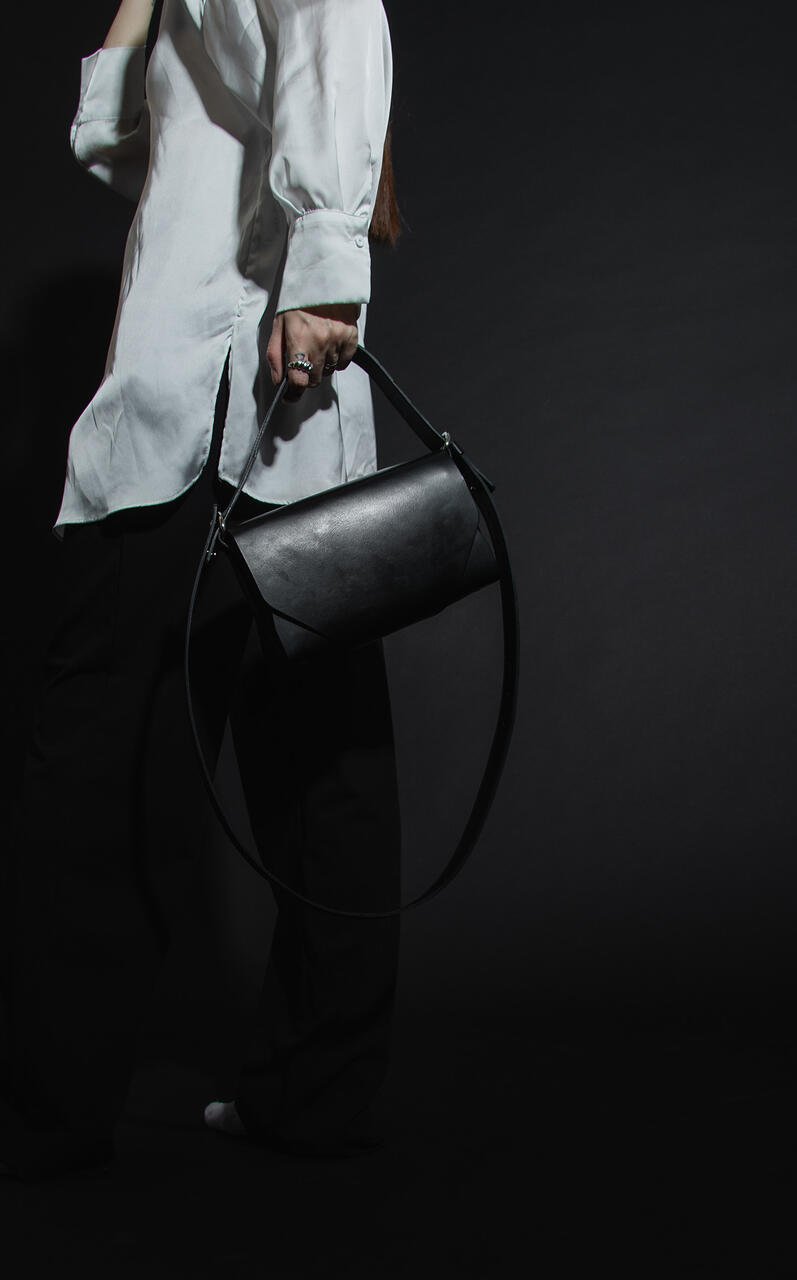 8. Woman holding black leather Stella Soomlais shoulder bag by strap, wearing white shirt and black pants in dark studio