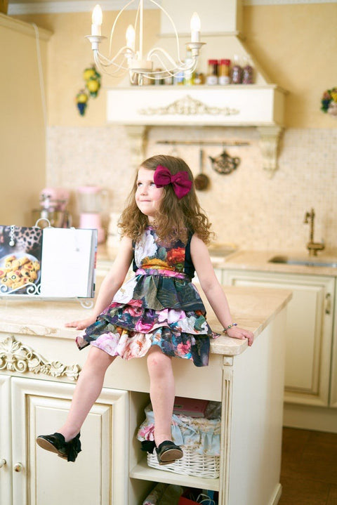 1. Young girl wearing Hortensias Home Francine Black kids apron with dark floral print, sitting on a kitchen counter, smiling, with a bow in her hair