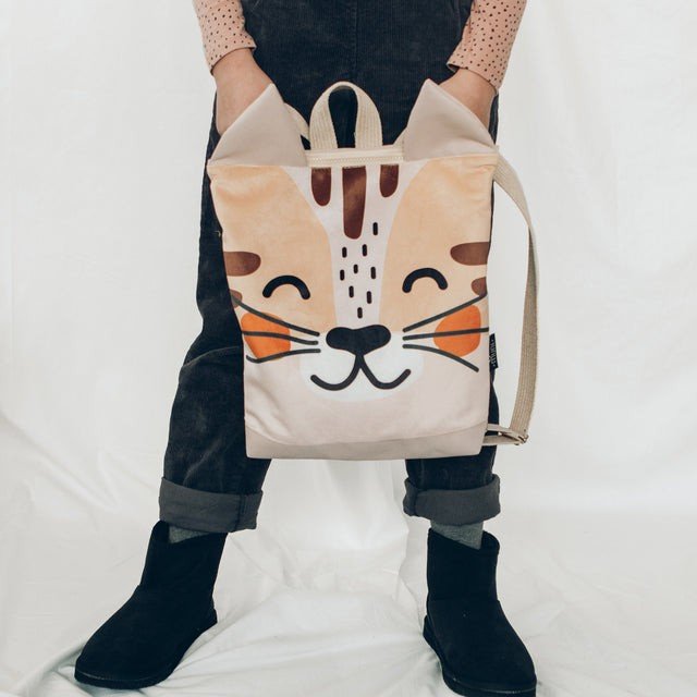 1. Child holding MUNI cat backpack by straps, wearing black pants and pink top, on white background