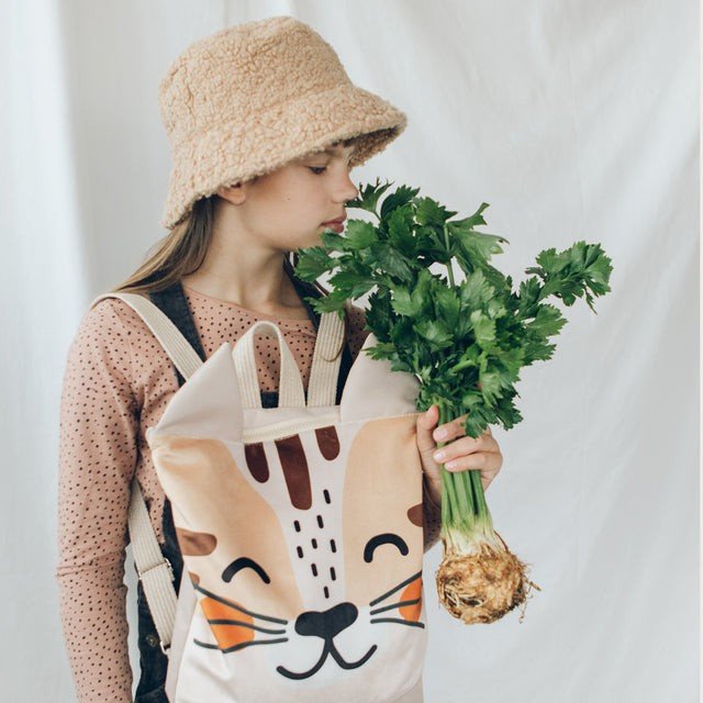 1. Girl with MUNI cat backpack and celery, wearing pink top and beige hat, on white background