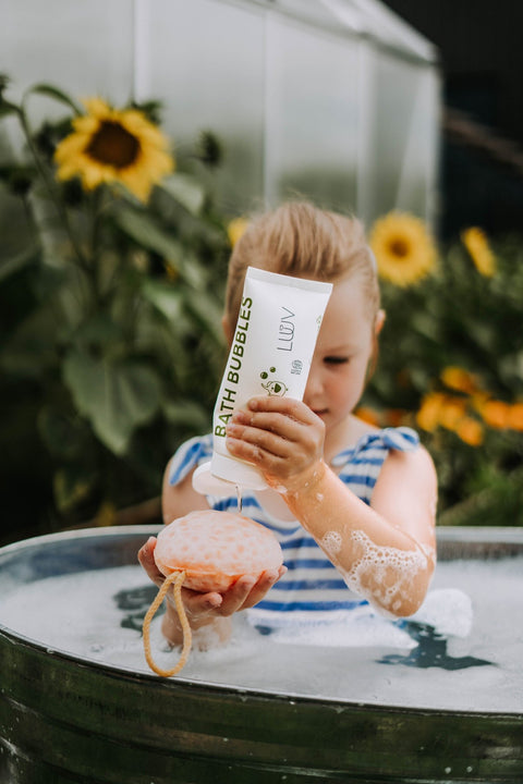 3. Child pouring LUUV pear-scented bath bubbles onto sponge in outdoor bath with sunflowers in background
