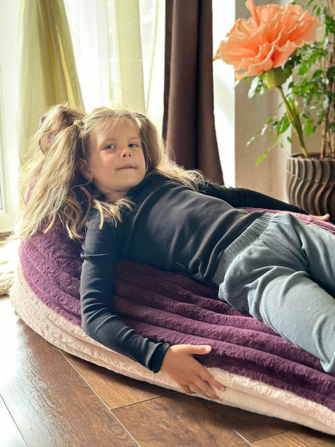 1. Girl relaxing on Luula kids bean bag chair in berry and rose mousse tones, in a bright room with curtains and a potted plant