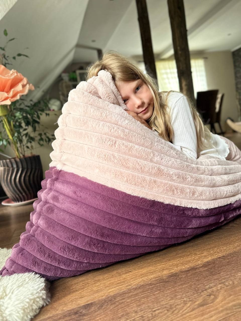 1. Girl resting on Luula kids bean bag chair in berry and rose mousse tones, in a cozy room with wooden beams and a large flower