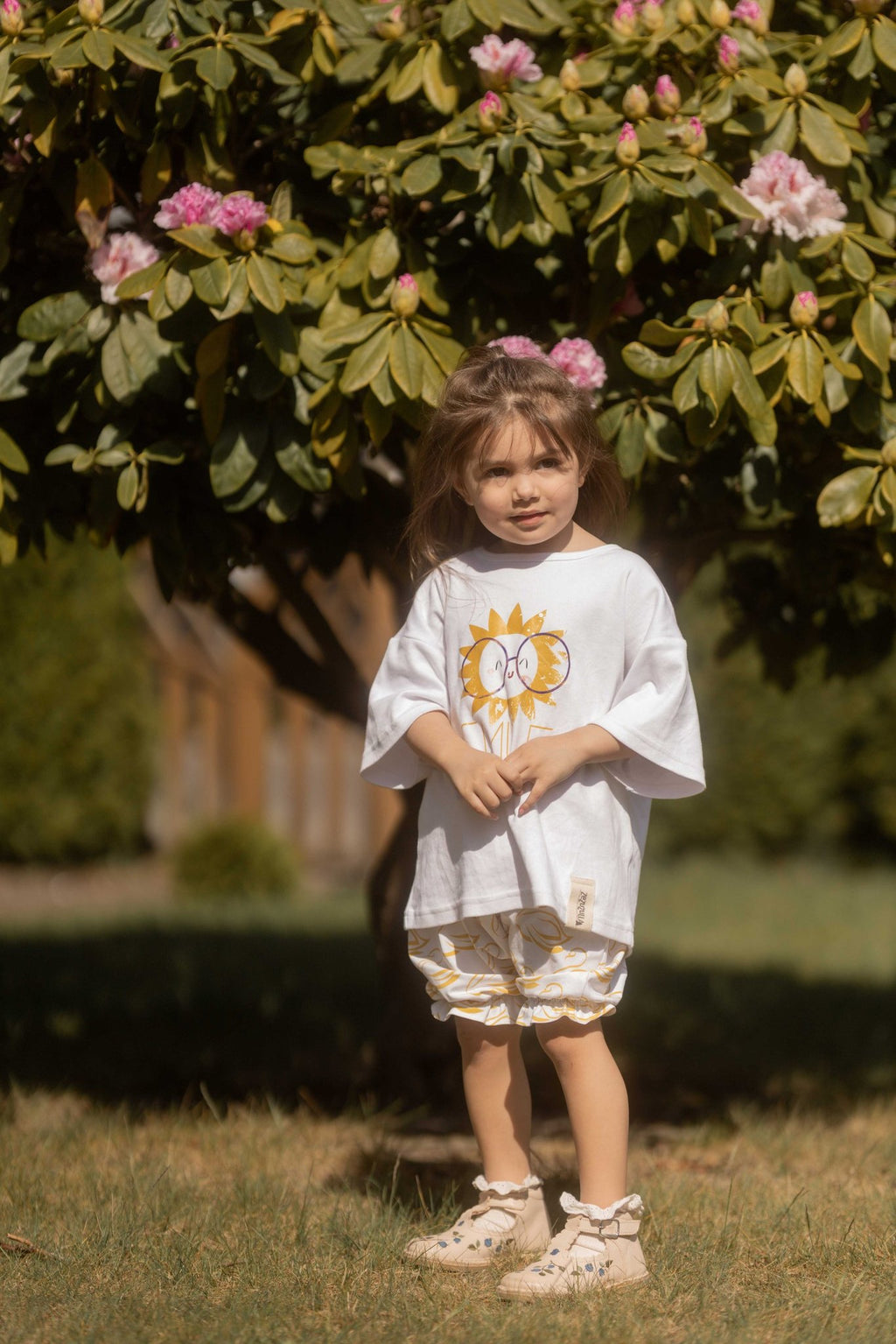 2. Young girl standing in garden wearing white bloomers with golden-yellow swans and oversized white t-shirt under a flowering tree