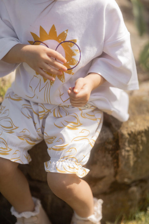 1. Child wearing white bloomers with golden-yellow swan print and ruffled leg openings, sitting outdoors on a stone