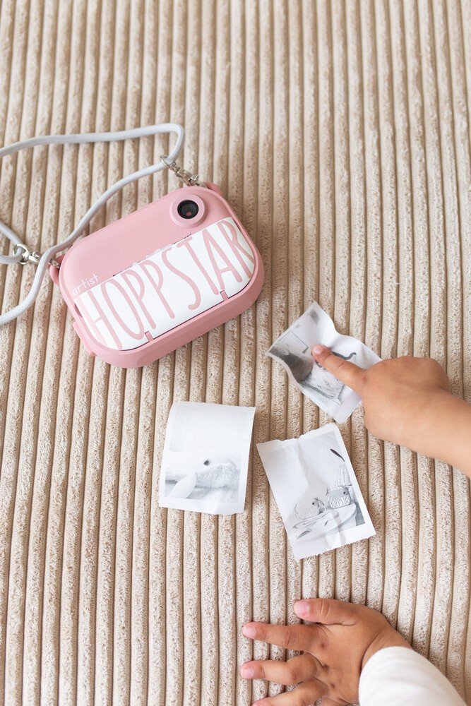 1. Child's hand holding pink Hoppstar camera with printed photos on striped fabric