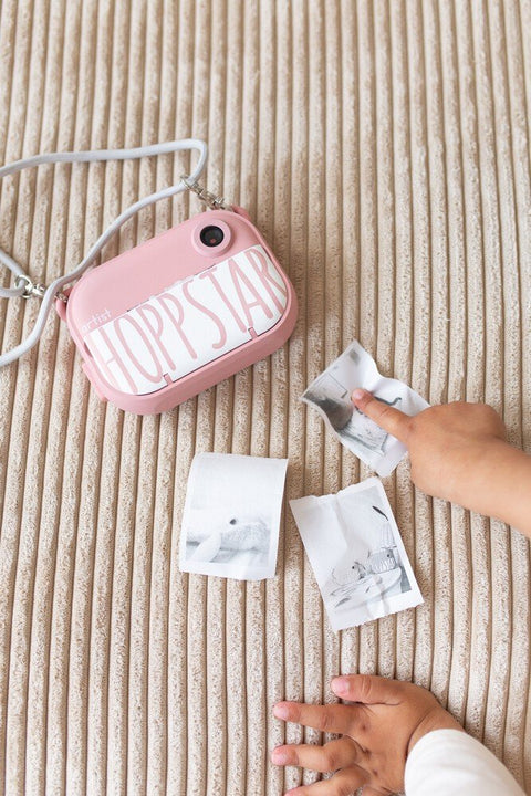 1. Child's hand holding pink Hoppstar camera with printed photos on striped fabric