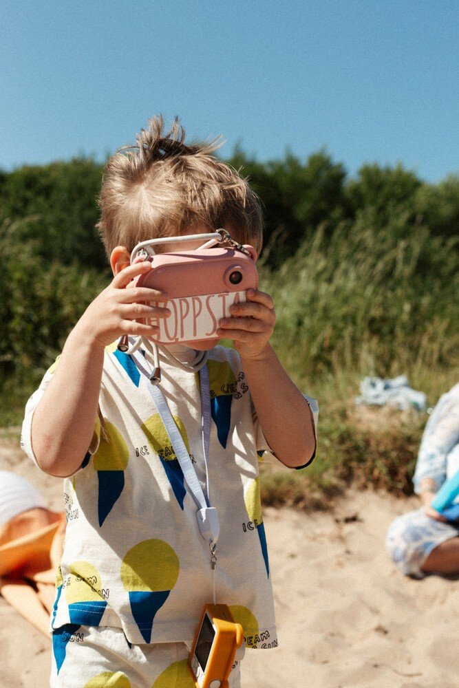 1. Child using pink Hoppstar camera on sandy beach with greenery in background