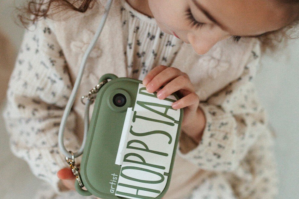 1. Child holding green Hoppstar camera with strap, wearing floral outfit indoors