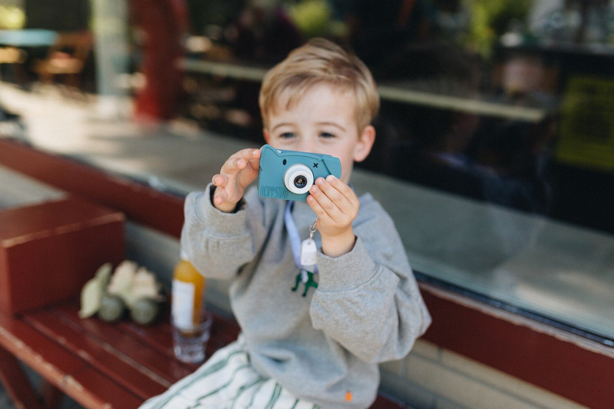 1. Boy using blue Hoppstar Rookie camera on bench, wearing casual outfit outdoors