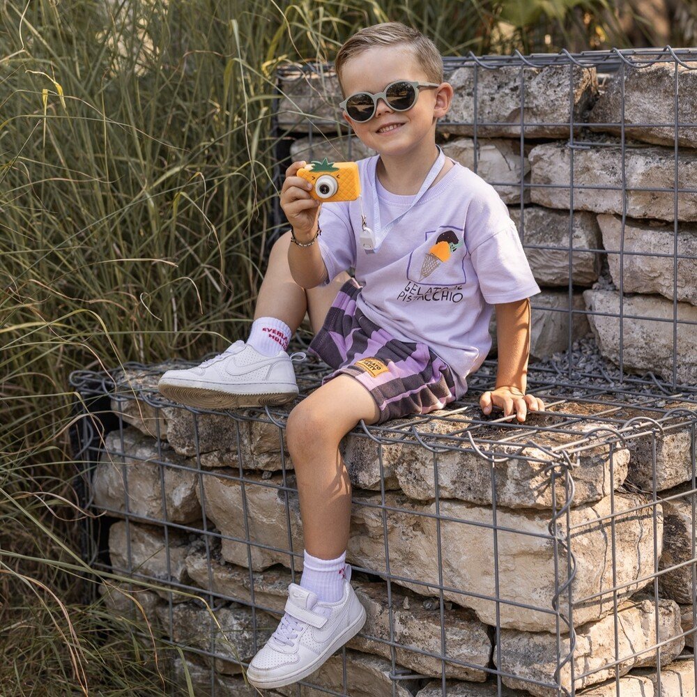 1. Boy sitting on stone wall holding yellow Hoppstar Rookie camera, wearing sunglasses and casual outfit outdoors
