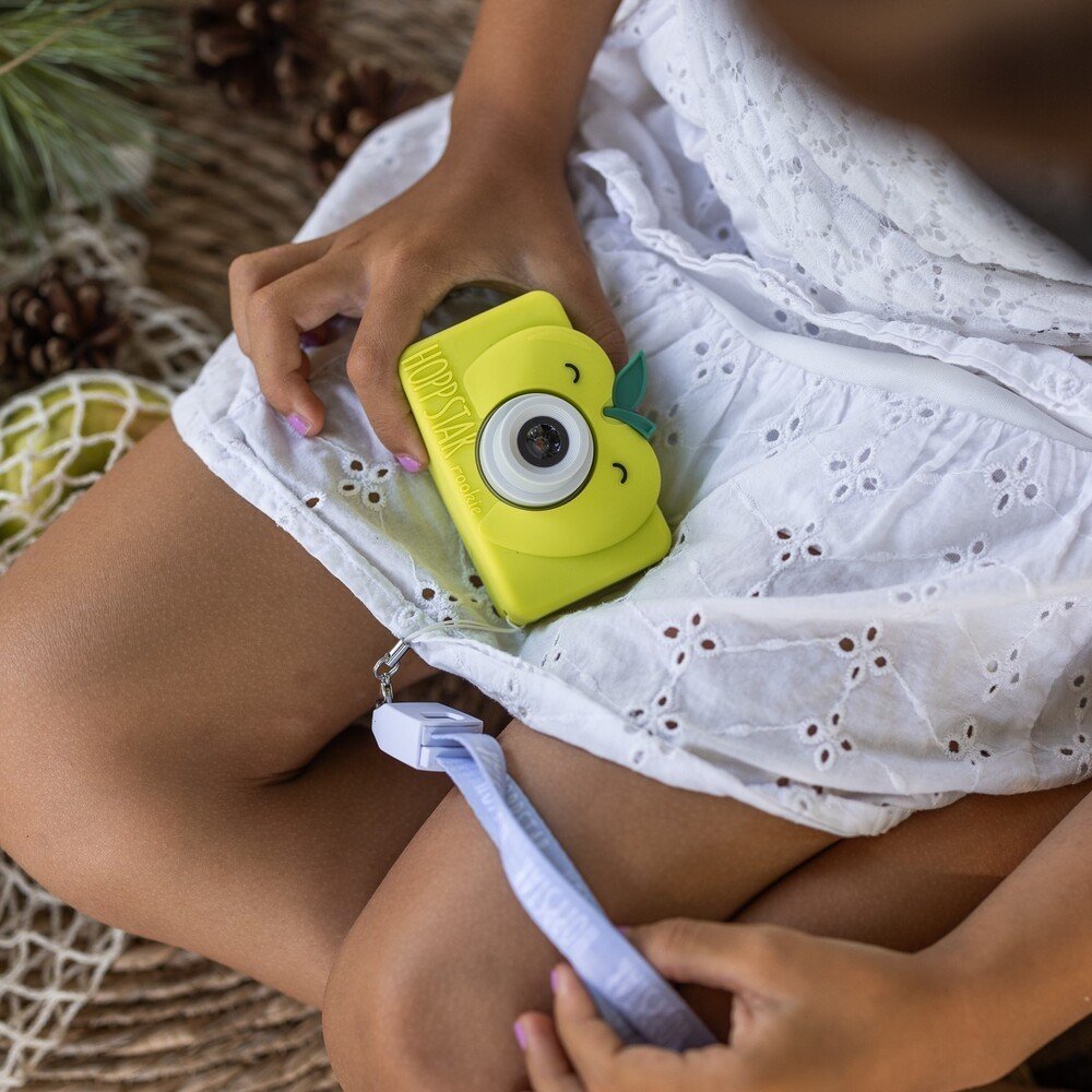1. Child holding green Hoppstar Rookie camera on lap, wearing white dress outdoors