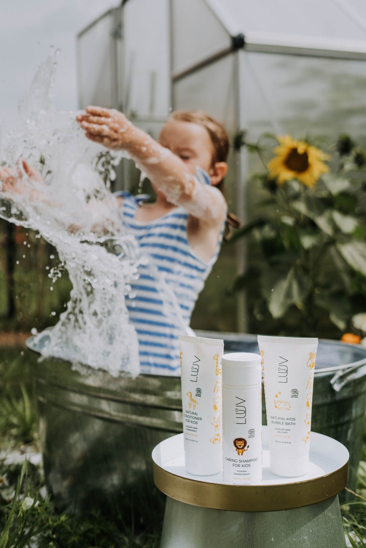 1. Child playing with water in garden setting with LUUV Kid's Conditioner Vanilla COSMOS and shampoo on table
