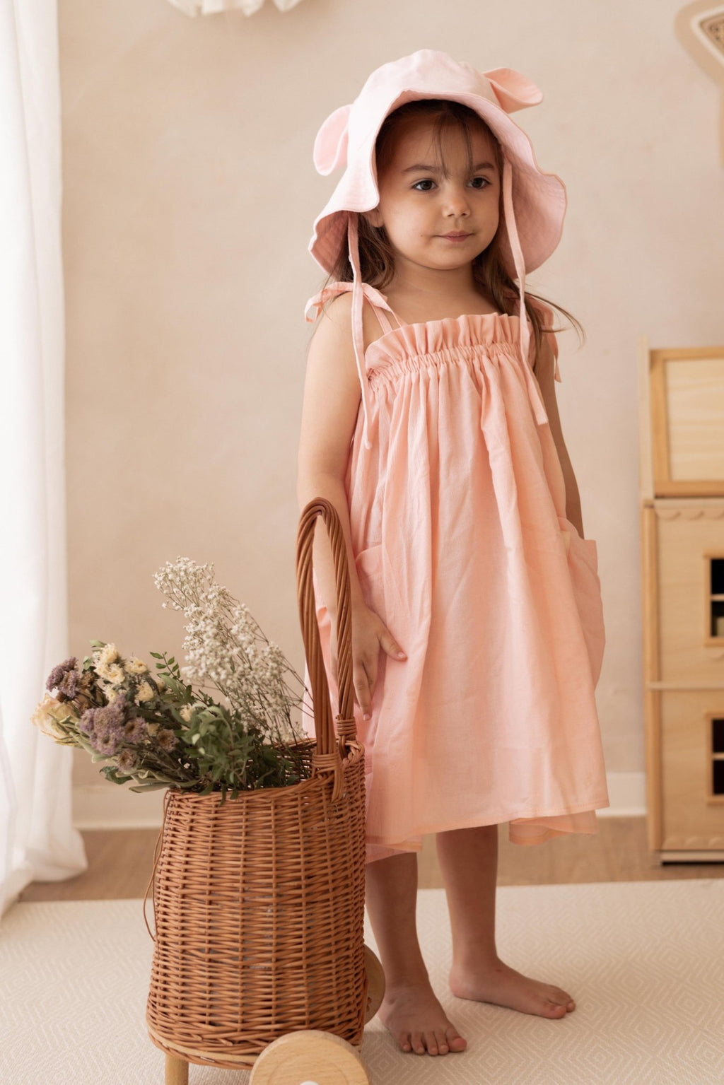 2. Girl in peach dress with pink hat standing next to wicker basket with flowers indoors