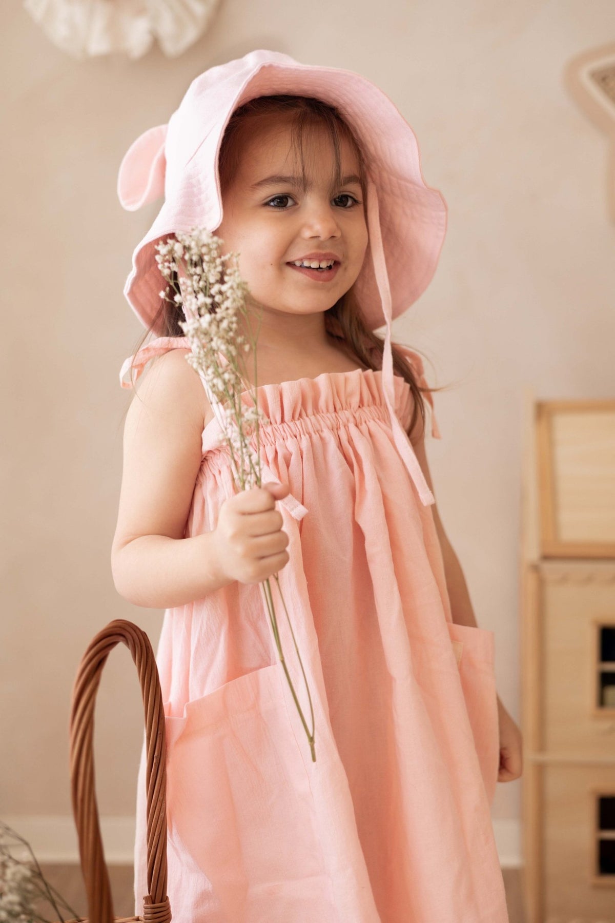 1. Girl wearing peach dress with adjustable shoulder ties and pink hat holding flowers indoors