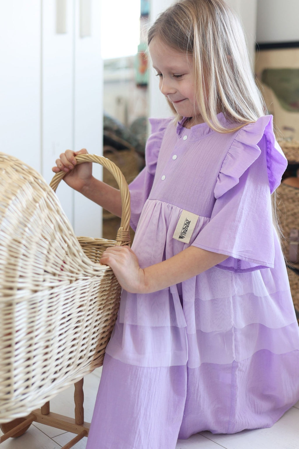 1. Girl wearing lilac Zezuzulla dress with ruffled sleeves and front snap buttons, playing with a wicker basket indoors