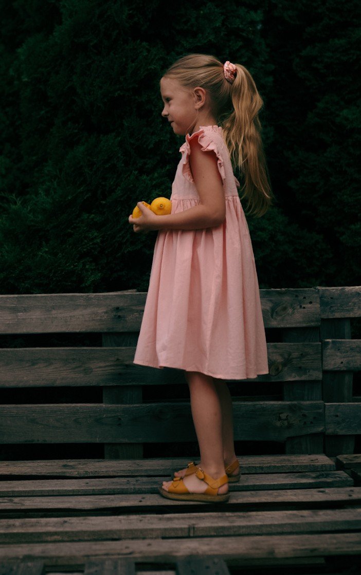 1. Girl wearing peach Zezuzulla dress with ruffled sleeves holding lemons, standing on wooden bench outdoors