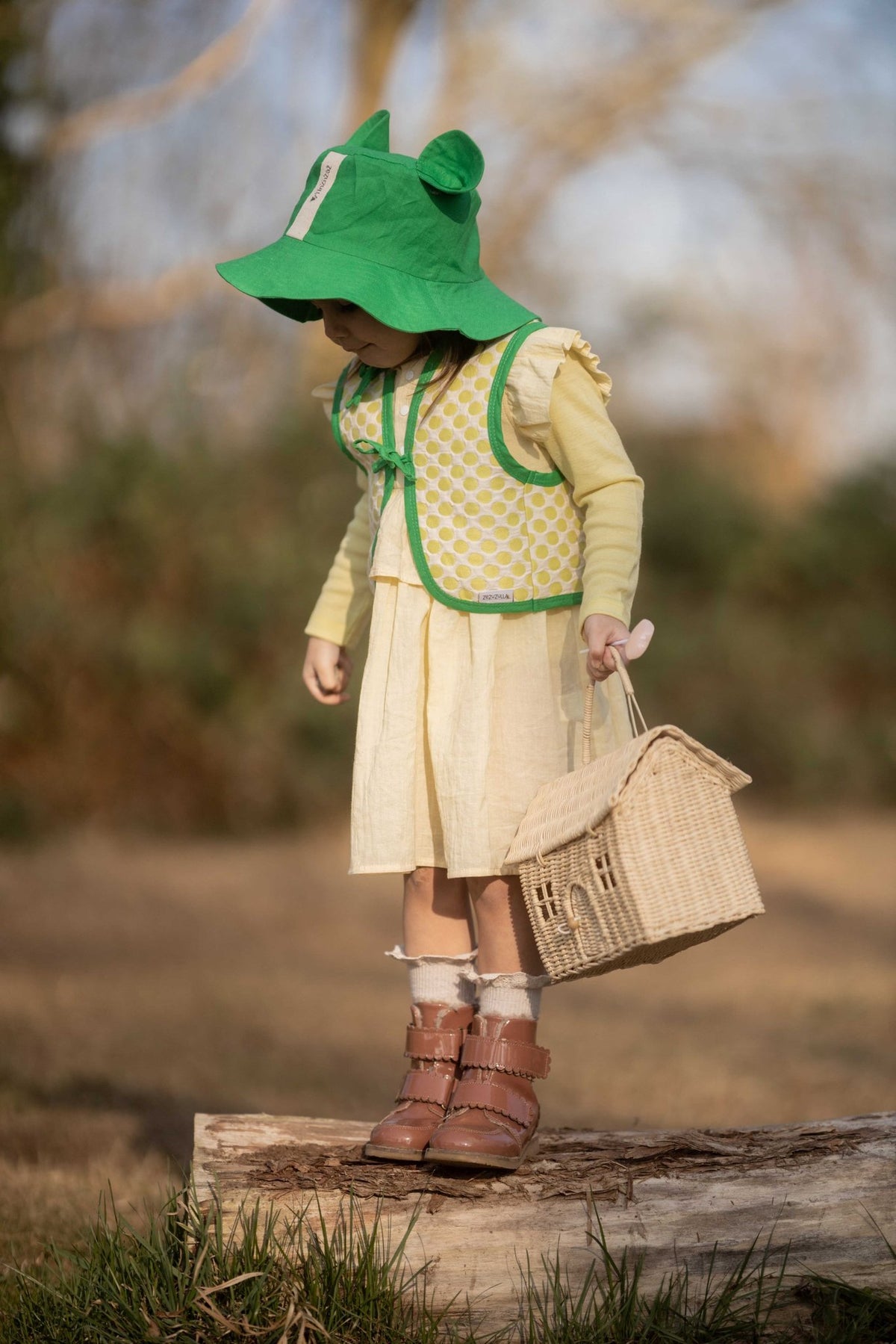 2. Child in yellow Zezuzulla dress with green hat and vest standing outdoors holding a basket