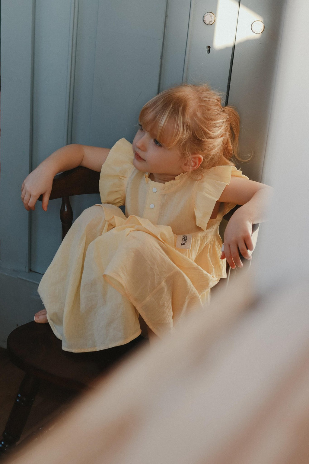 1. Young girl wearing yellow Zezuzulla dress with ruffled sleeves sitting on a chair indoors