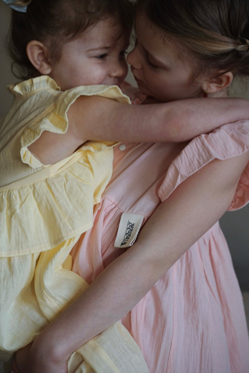6. Two children hugging, one in yellow Zezuzulla dress and the other in pink dress indoors