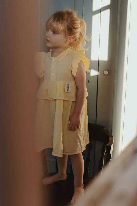 4. Young girl standing indoors wearing yellow Zezuzulla dress with ruffled sleeves and front buttons