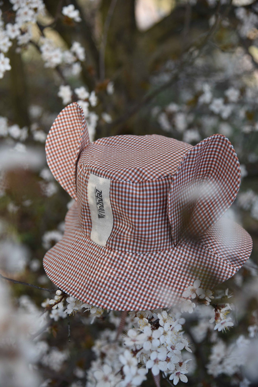 1. Zezuzulla kids chequered hat with fabric ears and brand label, placed among blooming branches in a natural setting