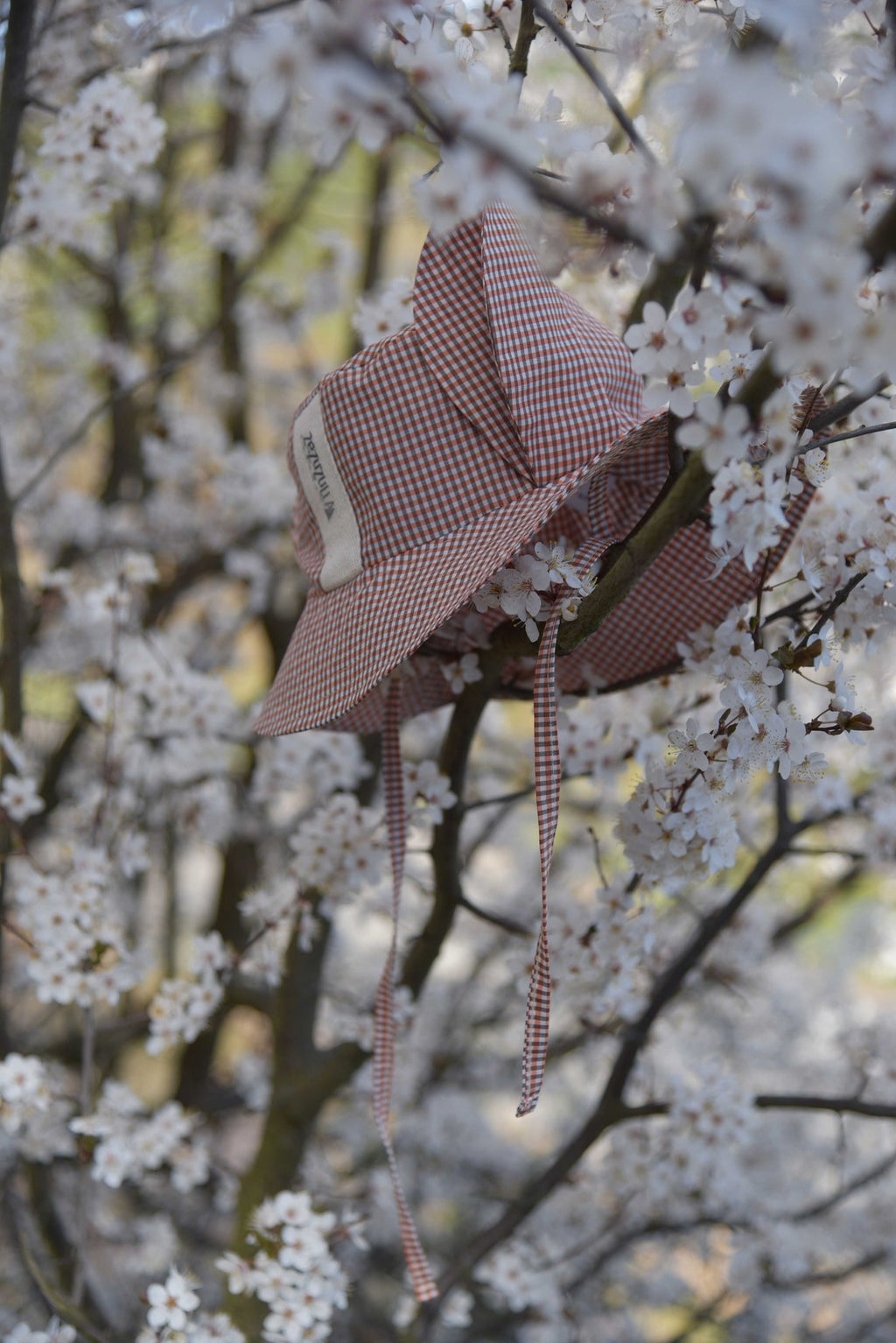 3. Side view of Zezuzulla kids chequered hat with fabric ears and ties, hanging on a tree branch with white blossoms