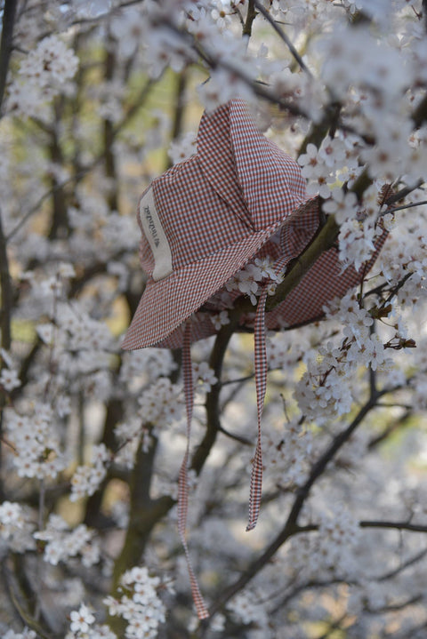 3. Side view of Zezuzulla kids chequered hat with fabric ears and ties, hanging on a tree branch with white blossoms
