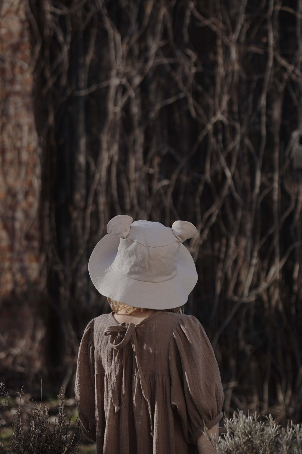 6. Back view of child wearing beige hat with ears in front of vine-covered wall