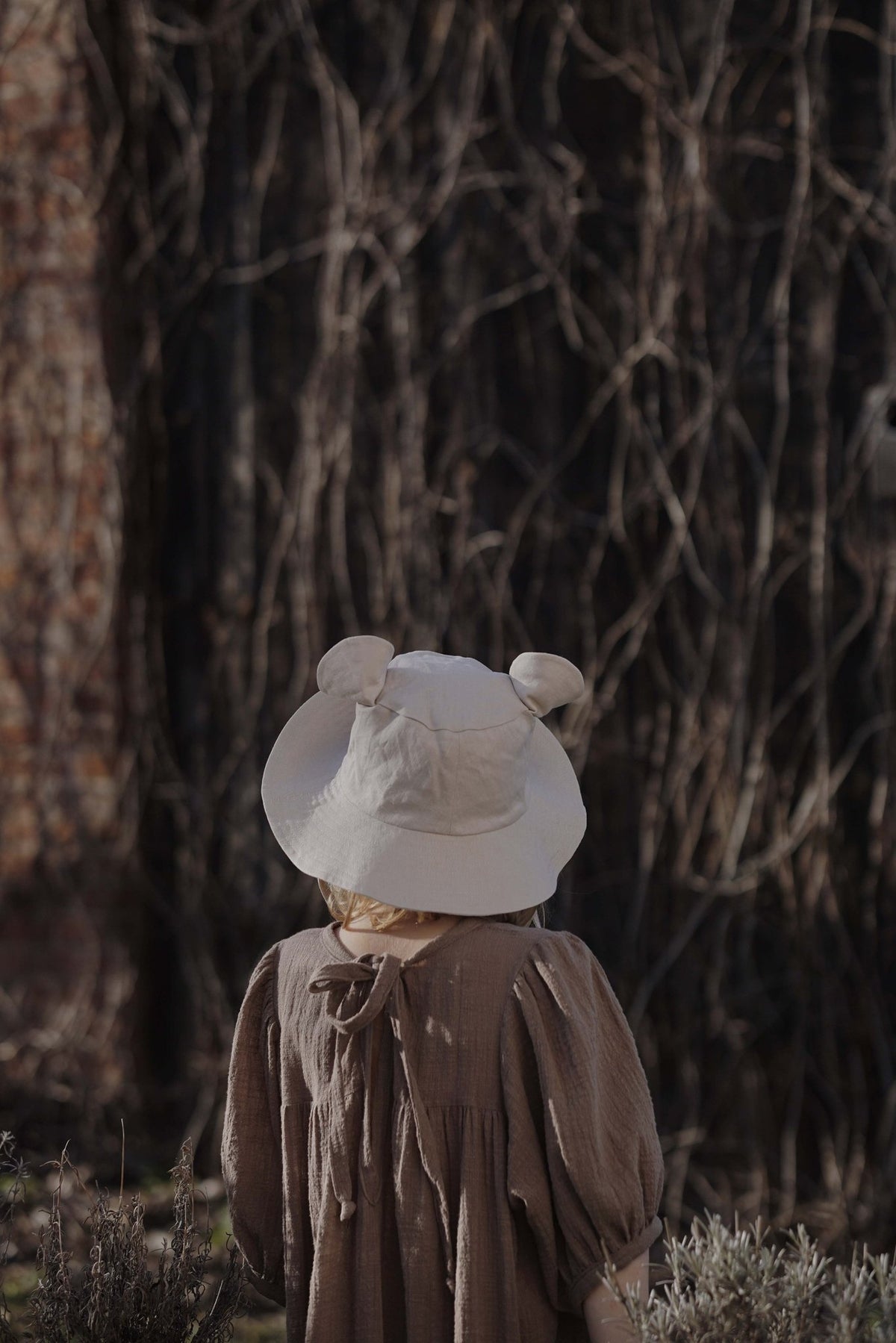 6. Back view of child wearing beige hat with ears in front of vine-covered wall