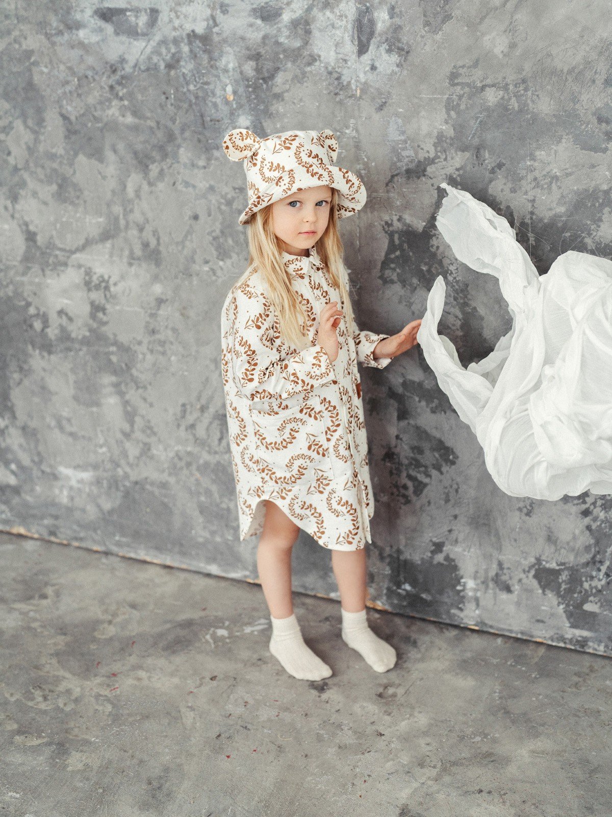 2. Child wearing Zezuzulla kids hat with ears and matching dress, standing against a textured grey wall