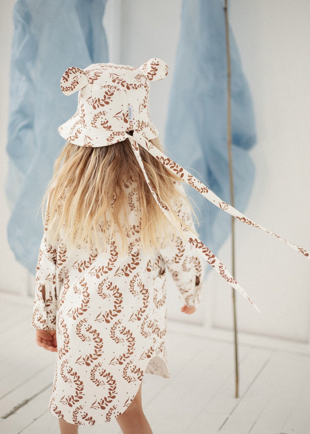 1. Child wearing Zezuzulla kids hat with ears and matching dress, shown from back in a bright room with blue fabric decor