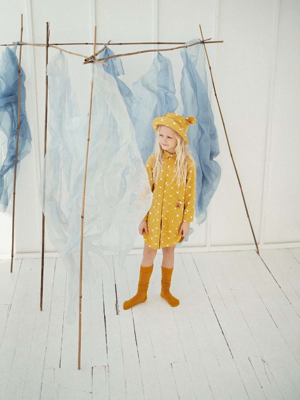 4. Child in mustard hat with ears and matching outfit, standing in white room with blue fabric decor