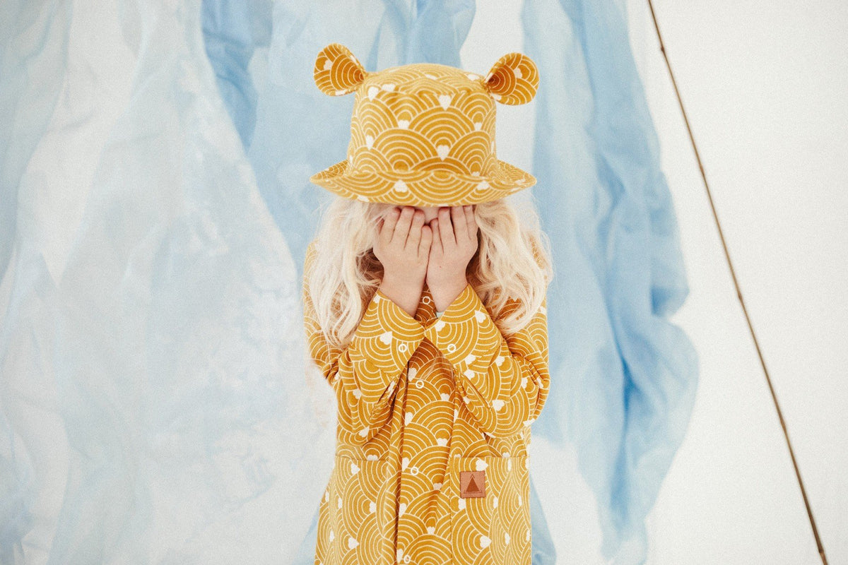 1. Child wearing mustard hat with ears and matching outfit, standing in front of blue fabric backdrop