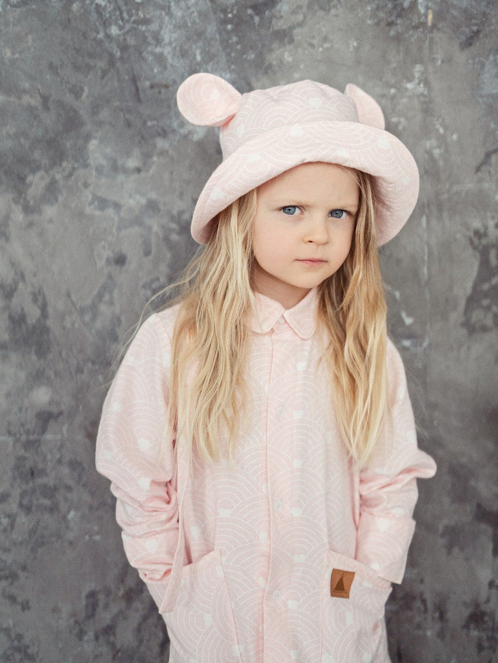 1. Child wearing pink hat with ears and wave pattern, standing against textured wall
