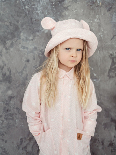 1. Child wearing pink hat with ears and wave pattern, standing against textured wall