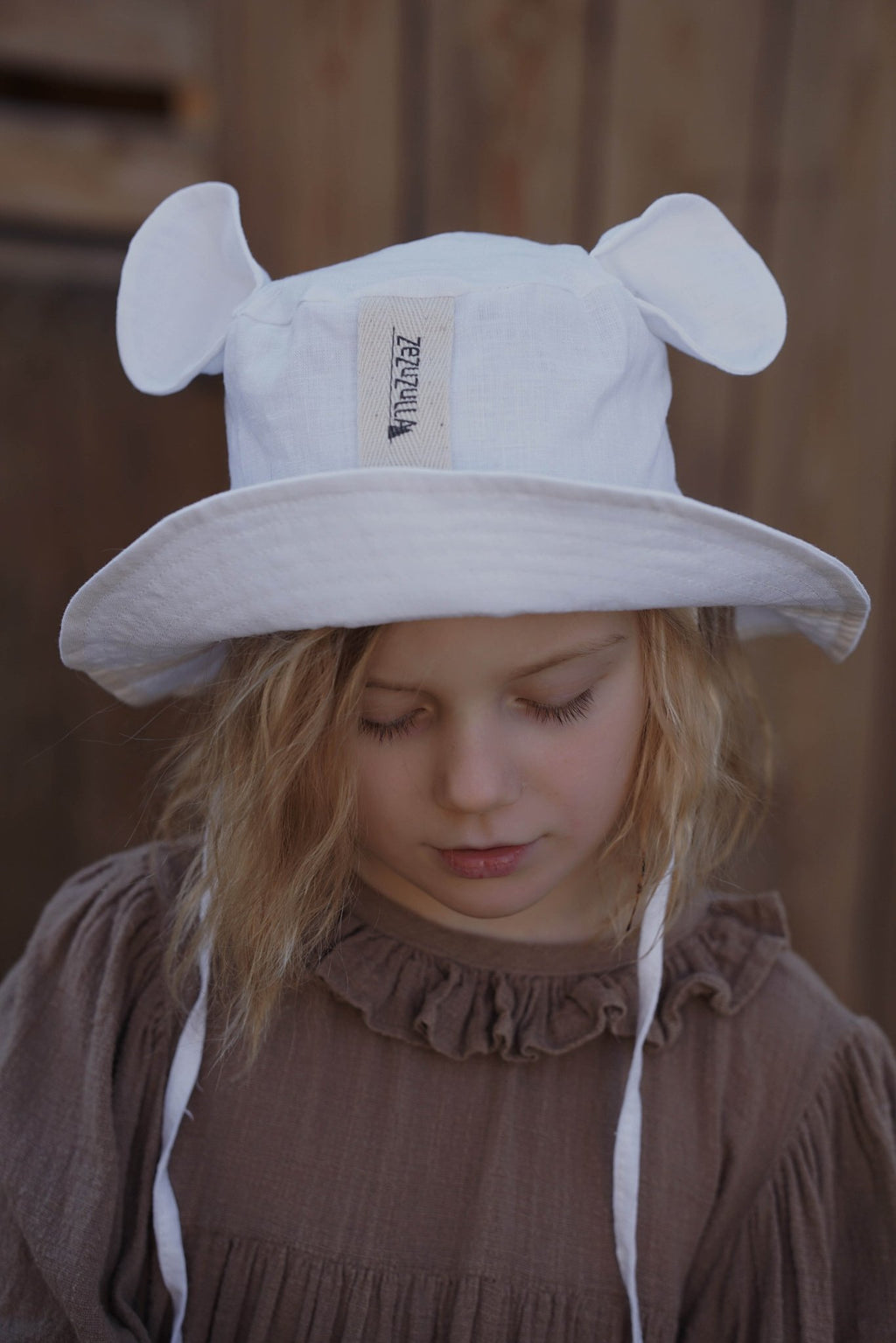 1. Child wearing white hat with ears and brown dress, looking down in outdoor setting