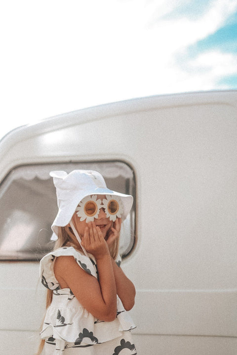 4. Child wearing white hat with ears and daisy sunglasses, standing in front of caravan