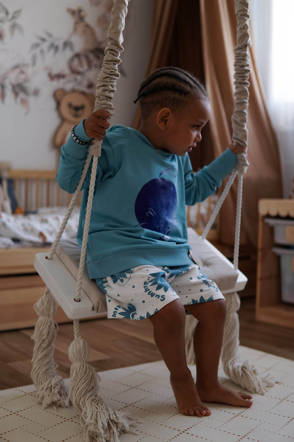 1. Child wearing blue hoodie with ears and moon print, sitting on a swing in a playroom setting