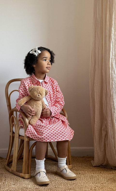 1. Young girl sitting on a chair holding a teddy bear, wearing Zezuzulla red patterned jacket and skirt set with white top