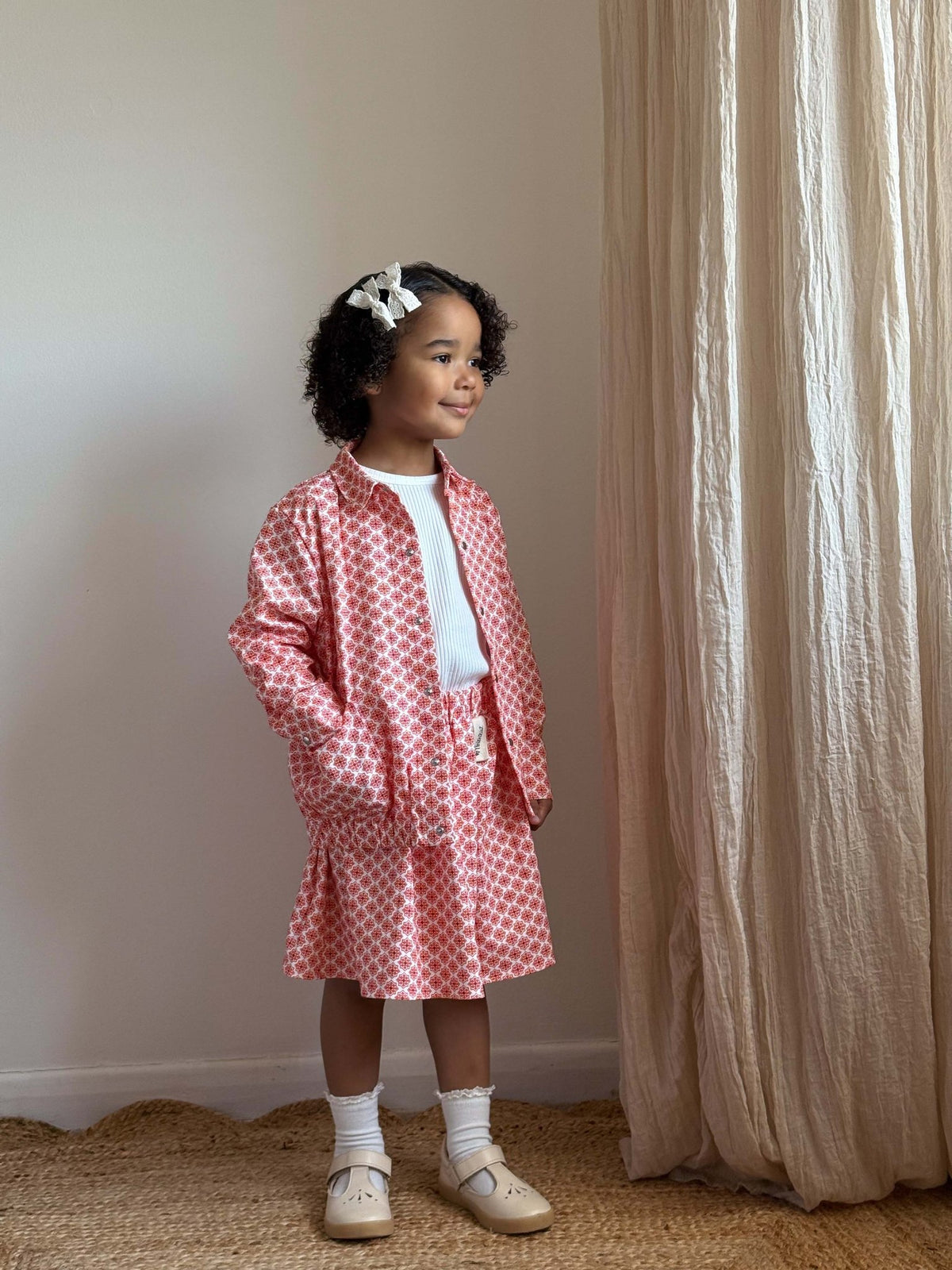 2. Full view of young girl in Zezuzulla red patterned jacket and skirt set with white top, standing in a softly lit room