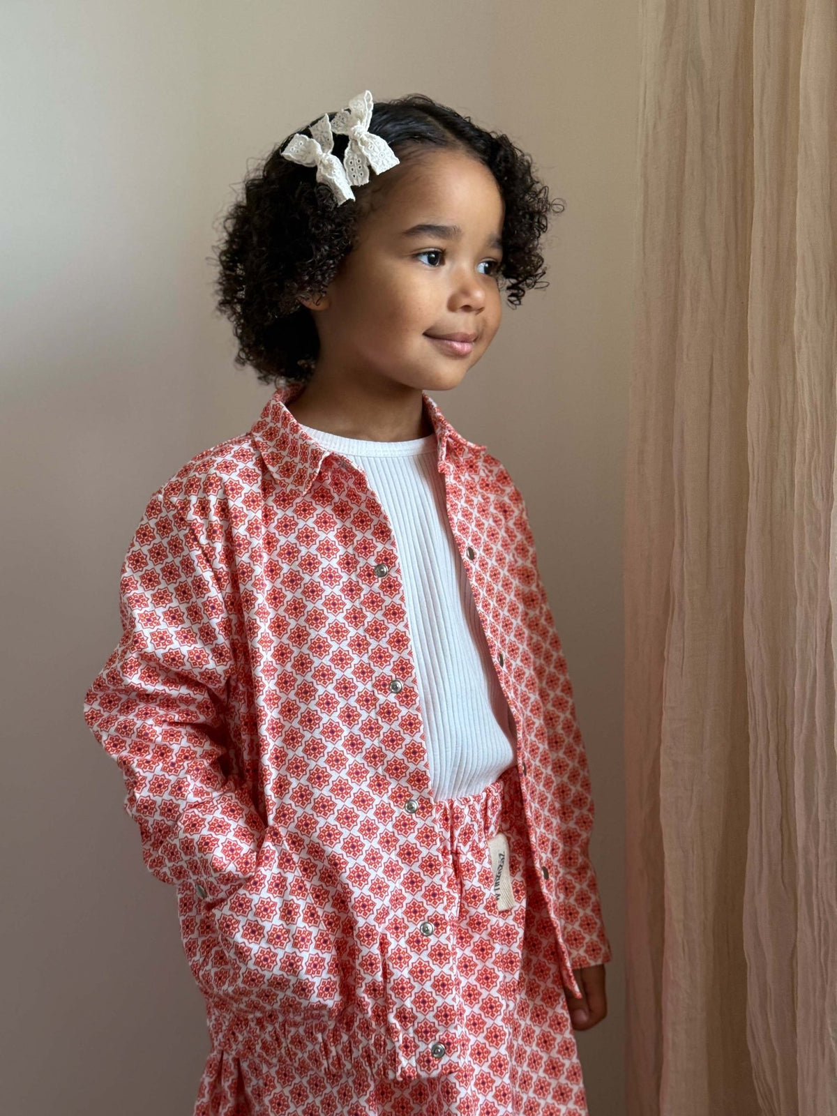 1. Young girl wearing Zezuzulla red patterned jacket and skirt set with white top, standing against a neutral background
